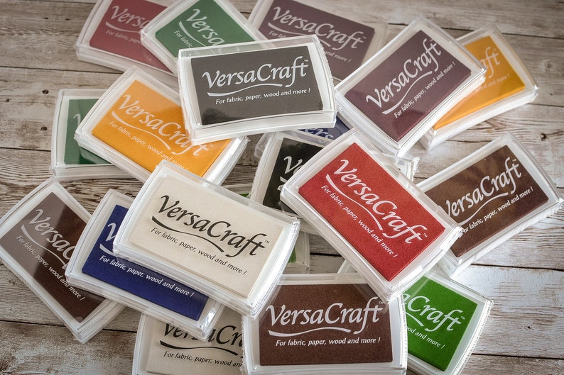 May include: A close-up of a pile of ink pads, all labeled "VersaCraft" and stating "For fabric, paper, wood and more!" The ink pads are in various colors, including brown, red, orange, white, and green.