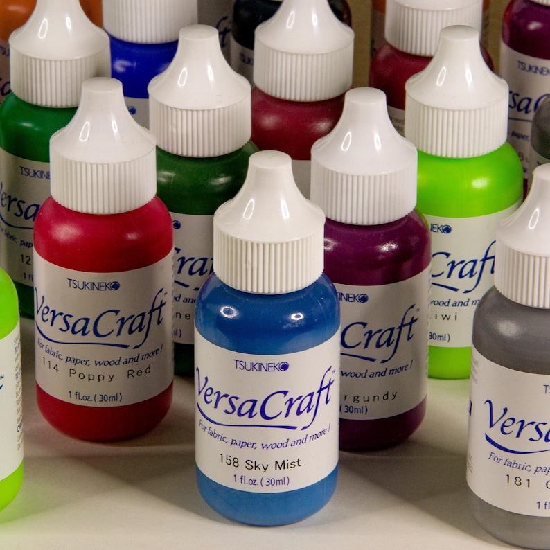 K&ouml;nnte beinhalten: Eine Sammlung von 10 kleinen Flaschen VersaCraft-Tinte in verschiedenen Farben, darunter Rot, Gr&uuml;n, Blau, Lila und Grau. Jede Flasche hat einen wei&szlig;en Verschluss und ein Etikett mit dem Markennamen und dem Farbnamen. Die Flaschen sind in einer Reihe auf einer wei&szlig;en Oberfl&auml;che angeordnet.
