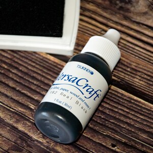 K&ouml;nnte beinhalten: Eine schwarze Flasche Tsukineko VersaCraft Tinte, beschriftet mit "Real Black" und "1 fl.oz. (30ml)". Die Flasche steht auf einer Holzoberfl&auml;che.