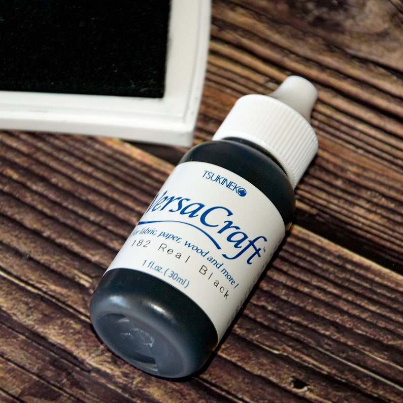 K&ouml;nnte beinhalten: Eine schwarze Flasche Tsukineko VersaCraft Tinte, beschriftet mit "Real Black" und "1 fl.oz. (30ml)". Die Flasche steht auf einer Holzoberfl&auml;che.