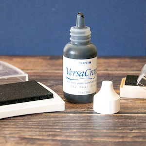 K&ouml;nnte beinhalten: Eine schwarze Tintenflasche mit der Aufschrift "VersaCraft" von Tsukineko. Die Flasche ist 1 Fl&uuml;ssigunze (30 ml) gro&szlig; und f&uuml;r Stoff, Papier, Holz und mehr geeignet.