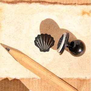 May include: A black seashell stamp and a black stamp with a circular design are displayed on a textured, light brown surface. A wooden pencil lies in the foreground. The stamps cast shadows.