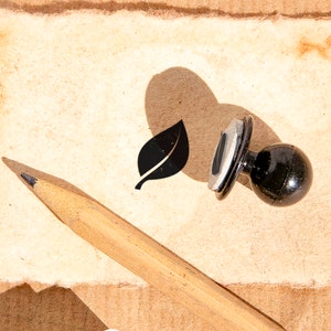 Könnte beinhalten: Ein schwarzer Gummistempel mit einem Blattmotiv auf braunem Papierhintergrund. Ein Bleistift aus Holz liegt auf dem Papier.