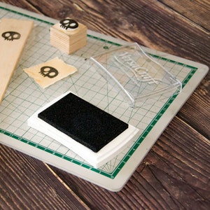 May include: A white ink pad with a black ink pad inside, a clear plastic container with the text "VersaCraft" on it, and a wooden block with a skull stamp on top. The items are on a green cutting mat.
