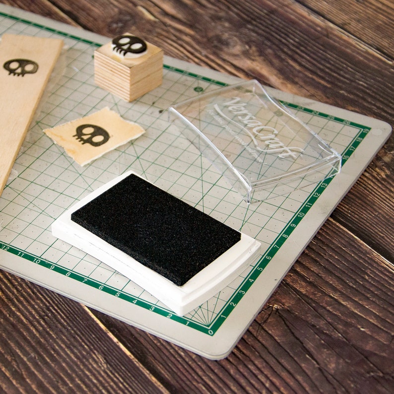 May include: A white ink pad with a black ink pad inside, a clear plastic container with the text "VersaCraft" on it, and a wooden block with a skull stamp on top. The items are on a green cutting mat.