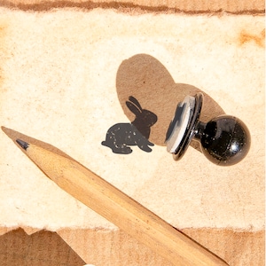 May include: A black rubber stamp with a rabbit design on a beige surface. A pencil is lying next to the stamp.