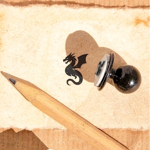 May include: A wooden pencil with a sharpened graphite tip lies near a black dragon stamp. The stamp has a round black handle and a silver metal stamp head. The dragon stamp casts a shadow on the textured, tan paper.