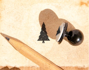 以下が含まれることがあります： 木製のハンドルが付いた黒いゴム製のスタンプで、クリスマスツリーの形をしています。このスタンプを使って、茶色の紙にクリスマスツリーの黒い印影を作っています。鉛筆が紙の上に置かれています。