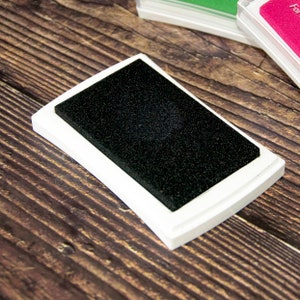 May include: A white ink pad with a black ink pad insert. The ink pad is rectangular and has rounded corners.