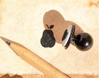 May include: A black rubber stamp with a pear design on a brown paper background. A wooden pencil is lying on the paper.