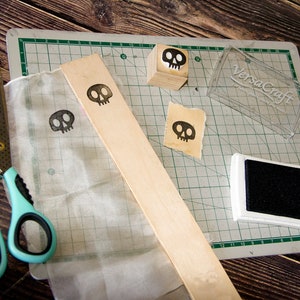 May include: A close-up of a craft project with a wooden block, a clear plastic container, a black ink pad, a piece of fabric, and a pair of scissors. The wooden block has a skull design on it. The clear plastic container has the text "VersaCraft" on it. The ink pad is black and rectangular. The fabric is white and has a skull design on it. The scissors are blue and silver.