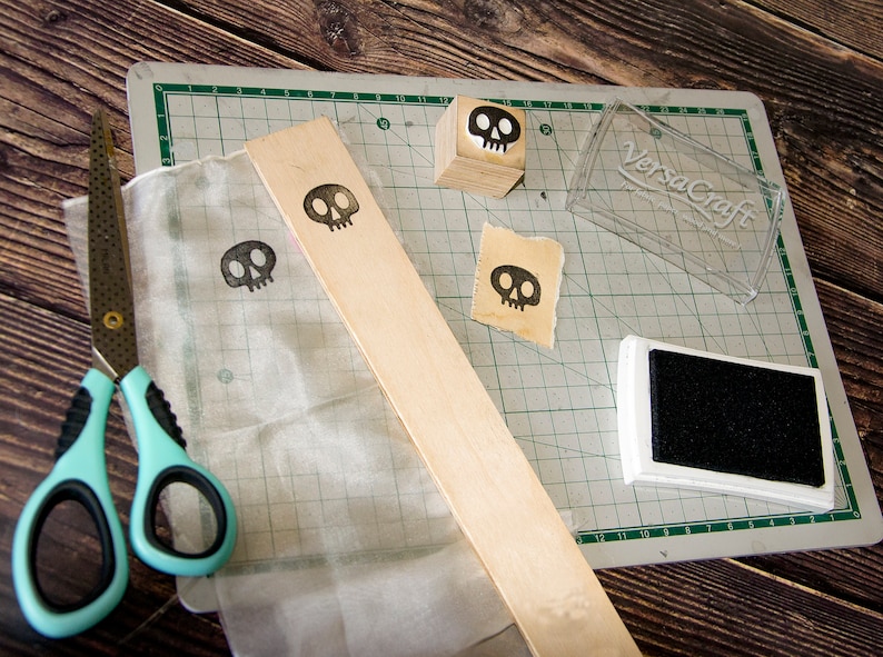 May include: A close-up of a craft project with a wooden block, a clear plastic container, a black ink pad, a piece of fabric, and a pair of scissors. The wooden block has a skull design on it. The clear plastic container has the text "VersaCraft" on it. The ink pad is black and rectangular. The fabric is white and has a skull design on it. The scissors are blue and silver.