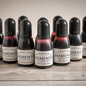 May include: A collection of ten small black bottles of Memento ink, each with a different color and name. The bottles are arranged in a row on a wooden surface. The ink is water-based, acid-free, and fade-resistant.