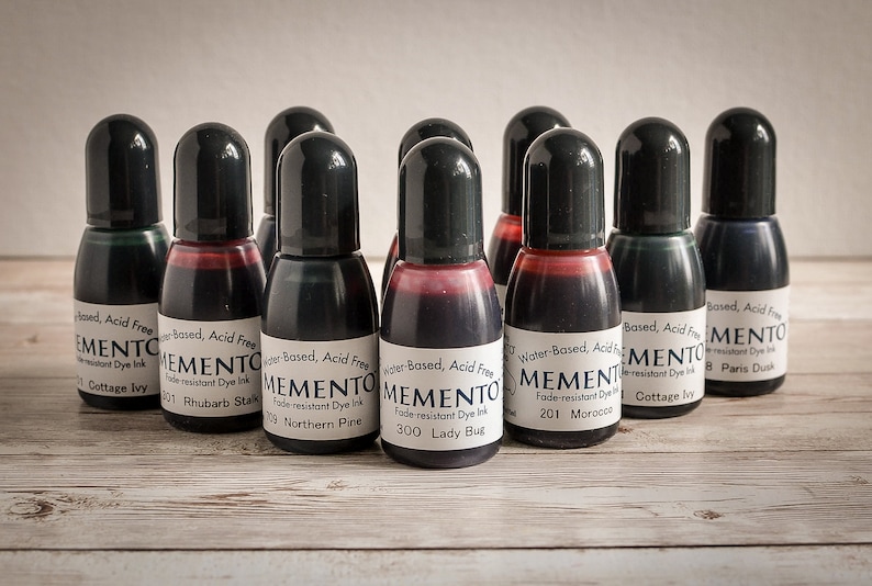 May include: A collection of ten small black bottles of Memento ink, each with a different color and name. The bottles are arranged in a row on a wooden surface. The ink is water-based, acid-free, and fade-resistant.