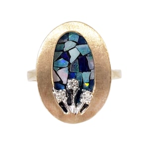 May include: Gold-toned ring with an oval face. The face features a mosaic of blue, teal, and white stones. Three small, silver-toned floral accents with clear stones are at the base of the oval.