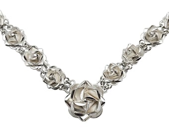 Könnte beinhalten: Eine silberne Halskette mit Rosenanhängern. Die Kette hat mehrere rosenförmige Anhänger, die durch eine feine Kette verbunden sind. Der zentrale Anhänger ist größer. Die Rosen haben ein detailliertes, dreidimensionales Design und eine polierte Oberfläche. Weißer Hintergrund.