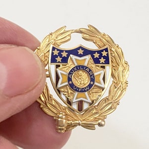 May include: Gold-toned pin with a blue and white shield featuring a yellow star and the words "AUXILIARY V.F.W." surrounded by a gold laurel wreath.