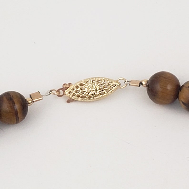 May include: A gold-toned clasp with a delicate filigree design connects two brown tiger's eye beads on a necklace.