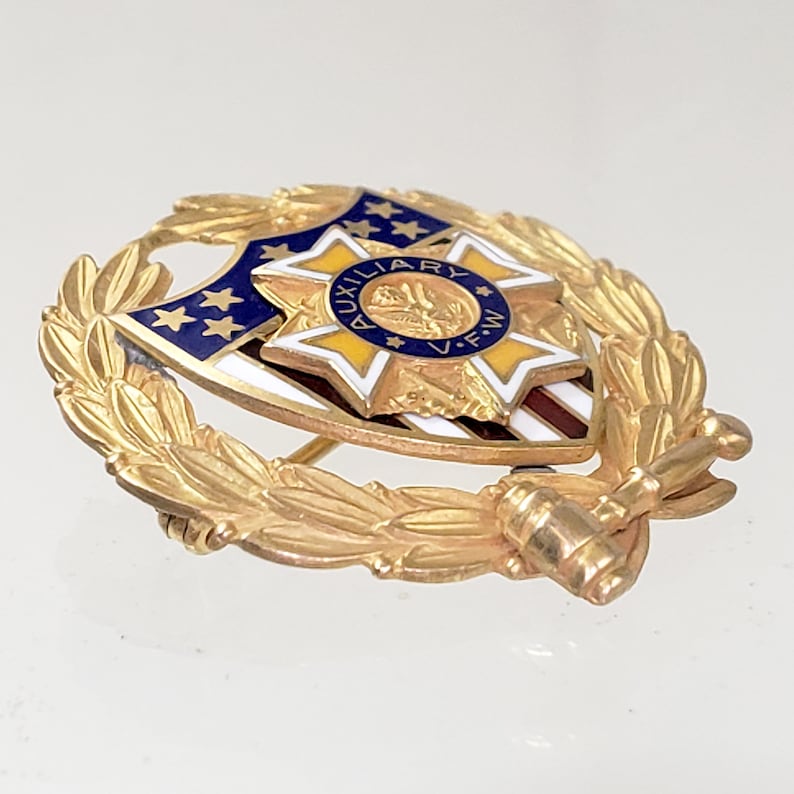 May include: Gold-toned pin with a blue and white shield featuring a five-pointed star with the words "AUXILIARY V.F.W." inside. The shield is surrounded by a wreath of gold leaves.