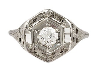 May include: A vintage-style ring with a central, round-cut diamond set in a geometric, filigree design. The ring's metal has a silver or platinum appearance, and the setting features intricate openwork details.