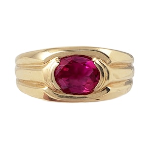 May include: A gold ring with a large, oval-shaped ruby gemstone set in the center. The band of the ring has three raised ridges.
