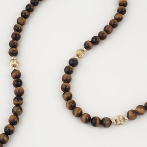 May include: A long brown tiger's eye beaded necklace with gold accents. The necklace is made of round beads and has a simple design.