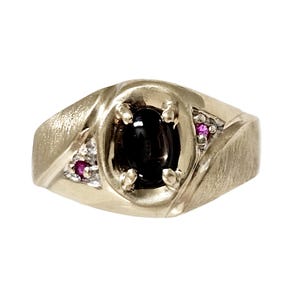 May include: A gold ring featuring a central oval black cabochon stone held by four prongs. The band has a brushed texture and is accented on each side by a triangular inlay containing a small, round, vibrant pink gemstone and a clear white stone.