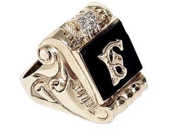 Könnte beinhalten: Ein goldfarbener Ring mit einem schwarzen Onyxstein und einem diamantbesetzten Oberteil. Der Ring hat ein verziertes Design mit einem stilisierten Buchstaben "B" auf dem schwarzen Stein. Der Ring ist wahrscheinlich ein Klassenring oder ein Siegelring.