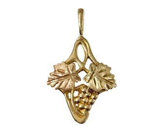 May include: A gold-colored pendant featuring a grape cluster and two leaves. The pendant has a bail for a chain and is set against a white background. The design is a stylized representation of a grapevine.