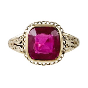 May include: A gold ring with a large, square-shaped, deep pink gemstone set in a decorative, gold frame.