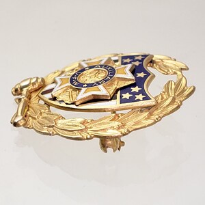 May include: Gold-toned pin with a blue and white enamel shield featuring a star and the text "AUXILIARY F.W.". The shield is surrounded by a gold laurel wreath.
