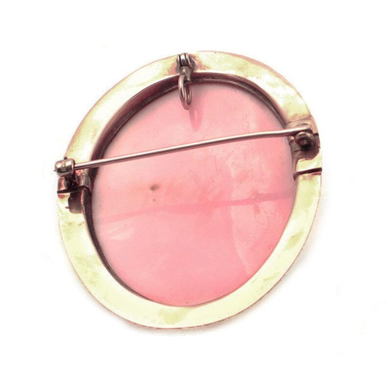 May include: A pink cameo brooch with a gold-toned metal setting. The brooch has a pin and a loop for attaching to clothing.