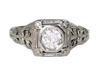 May include: A silver filigree ring with a single round diamond set in a square bezel. The ring has a delicate floral design.