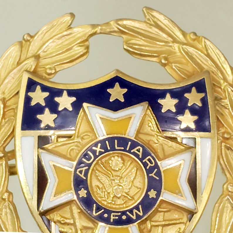 May include: Gold-toned pin with a blue and white shield featuring five gold stars and the text "AUXILIARY V.F.W."