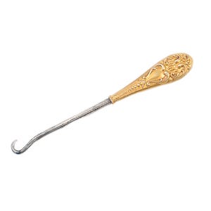 May include: A gold and silver shoe horn with an ornate handle. The handle is decorated with a floral design.