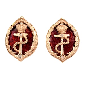 May include: Pair of gold-toned oval-shaped earrings. Each earring features a red enamel background with a gold anchor and crown design. The anchor is intertwined with a rope, and the crown is at the top. The earrings are framed by a gold laurel wreath.