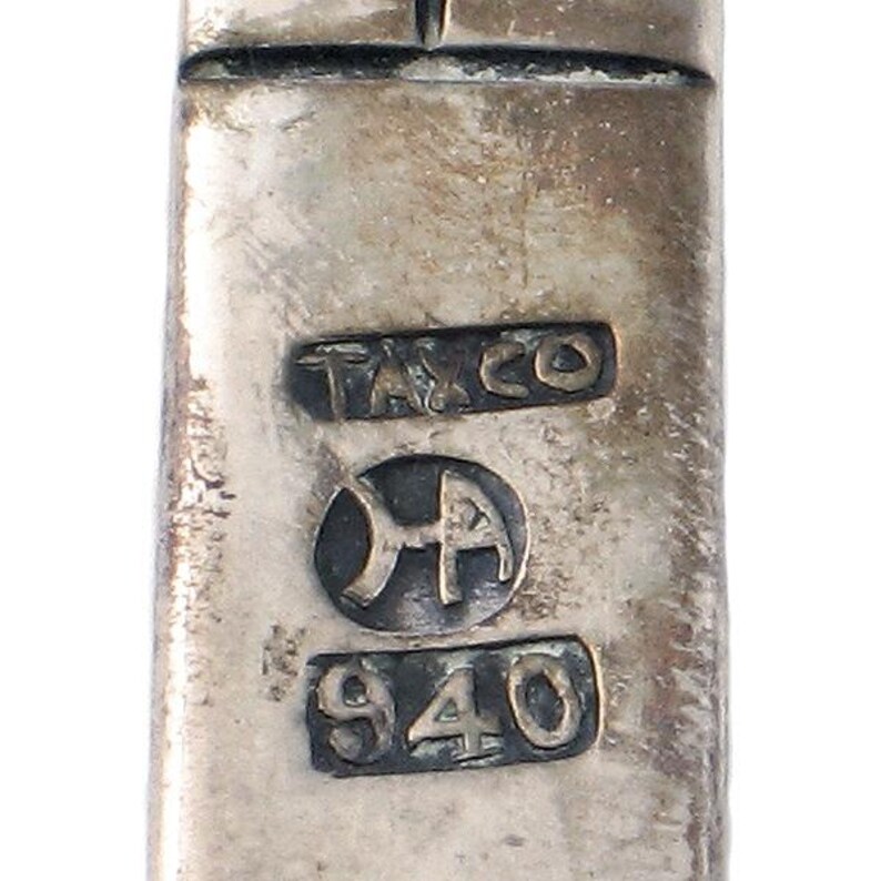K&ouml;nnte beinhalten: Nahaufnahme eines silberfarbenen Metallobjekts mit den Markierungen "T&CO" und "940" sowie einem Symbol, das wie ein stilisiertes "A" aussieht.