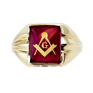 May include: A gold-colored ring with a rectangular, deep red stone. The stone features a gold-colored Masonic symbol. The ring has a ribbed design on the sides and a polished finish, showcasing a classic and elegant style.