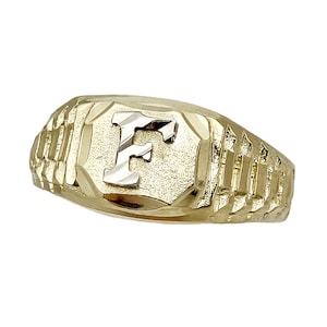 May include: A gold-toned ring with a textured band and a rectangular face. The face features the letter "F" in a contrasting silver color. The ring is likely made of gold and is designed for wear on a finger.