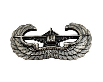 Op de afbeelding: Een zilverkleurige militaire badge met een gestileerd vliegtuigontwerp met uitgespreide vleugels. De badge heeft ingewikkelde details, waaronder veerachtige texturen op de vleugels en een gedetailleerd vliegtuigsilhouet. Waarschijnlijk een militair insigne.