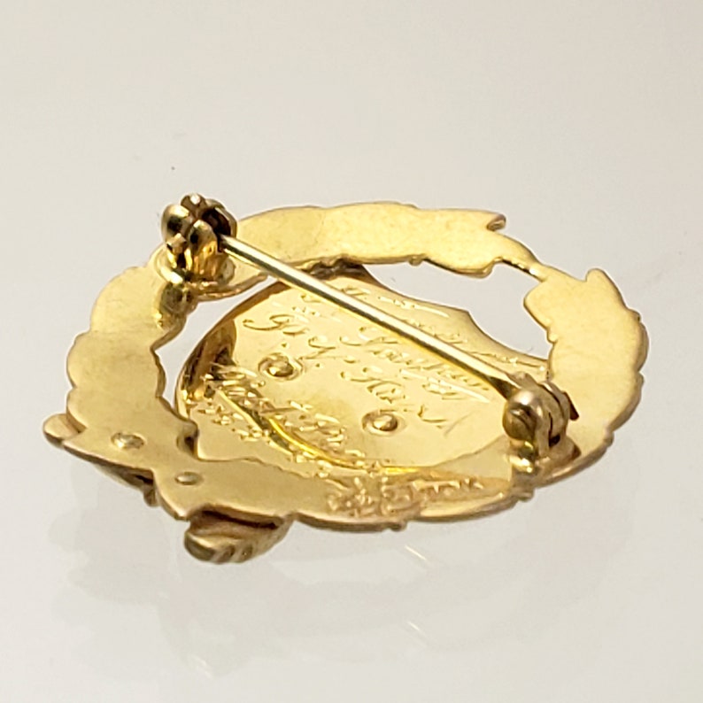 May include: Gold-toned pin with a wreath design and a circular center with engraved text. The pin has a safety clasp on the back.