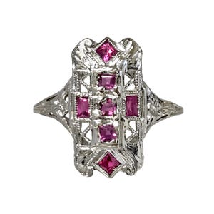 May include: A silver filigree ring with a rectangular design. The ring features five rectangular and two square, bright pink gemstones. The ring has an ornate, openwork design with floral accents.