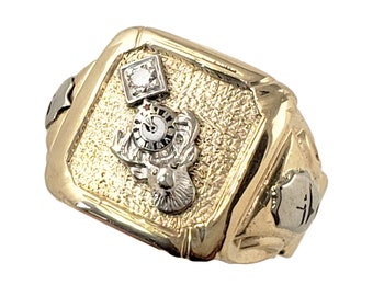 May include: A gold ring with a square face. It features a diamond and a detailed emblem of a ram's head and a clock. The ring has a textured surface and a silver accent on the side.