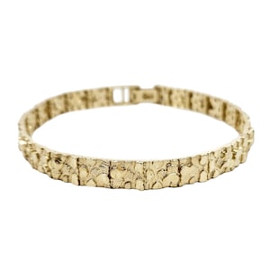 May include: A gold-colored nugget bracelet with a textured surface. The bracelet is made of individual rectangular links connected together. The clasp is visible at the top of the bracelet.