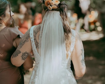 Op de afbeelding: Een witte tule bruidssluier bevestigd aan een haaraccessoire met een cluster van oranje, perzik- en crèmekleurige bloemen met groen blad. De sluier is doorschijnend en valt over de rug, gecombineerd met een mouwloze witte kanten jurk.