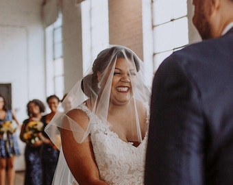 May include: A person wearing a white lace wedding dress with a V-neckline and a sheer tulle veil covering their head and face. The person is smiling broadly while facing another individual wearing a dark blue suit jacket during a ceremony.