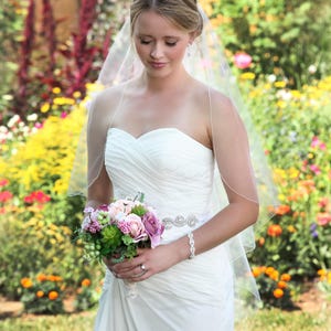 May include: A white strapless wedding dress with a ruched bodice and a jeweled belt, paired with a sheer, layered tulle veil. The ensemble includes a silver-toned bracelet and a bouquet of pink, purple, and green flowers. Text reads Bliss Veils and Accessories.