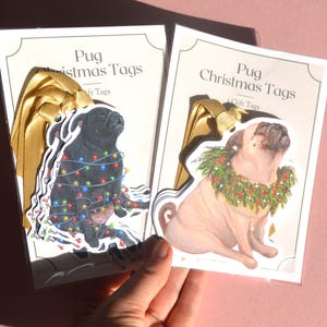 May include: Two sets of Pug Christmas Tags in clear packaging. One set features a black pug wrapped in colourful Christmas lights, the other a fawn pug wearing a green holly wreath. Both tags include gold ribbon loops and printed backing cards.