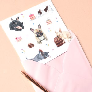 May include: A white greeting card featuring a pattern of French bulldogs in various colors interacting with slices of cake and cupcakes, partially tucked into a light pink envelope with a metallic copper pen resting alongside the stationery.