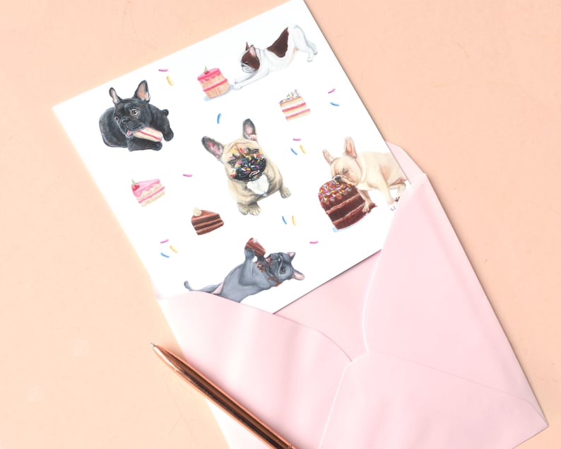May include: A white greeting card featuring a pattern of French bulldogs in various colors interacting with slices of cake and cupcakes, partially tucked into a light pink envelope with a metallic copper pen resting alongside the stationery.